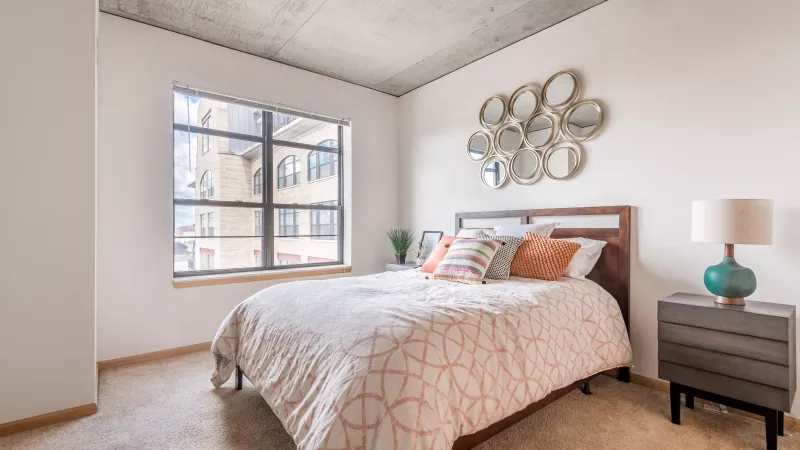 Modern bedroom with a bed, orange patterned comforter, wood headboard, large window, concrete ceiling, and decorative mirrors.