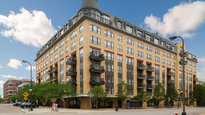 Tan multi-story building with dark balconies and a distinctive turreted roof, set against a blue sky.