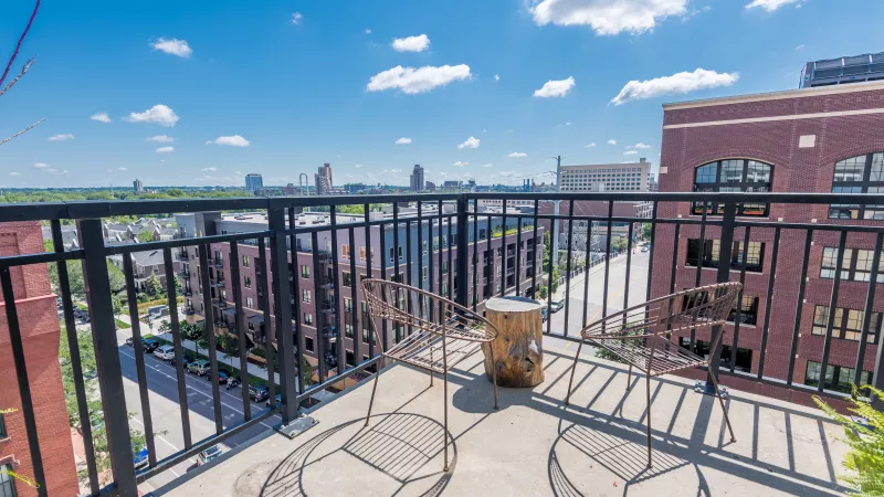 Rooftop balcony with two metal chairs, a barrel table, black railing, and a city skyline under a blue sky.