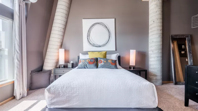 Modern bedroom with a white bed, gray walls, and exposed concrete pillars. Abstract art, warm lamps, and a tall mirror.