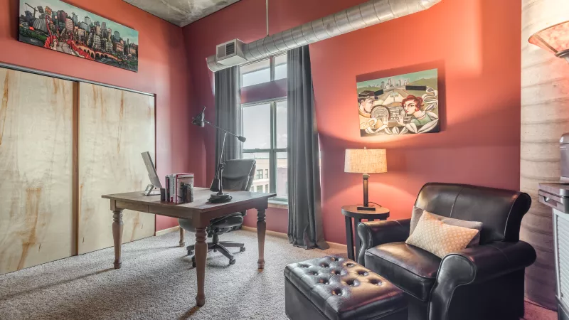 Cozy office with reddish walls, a wooden desk, black leather armchair with ottoman, and exposed industrial ductwork.
