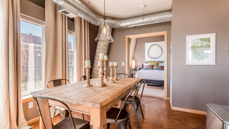 Dining area with a rustic wooden table, industrial chairs, exposed ducts, and an open doorway to a bedroom.