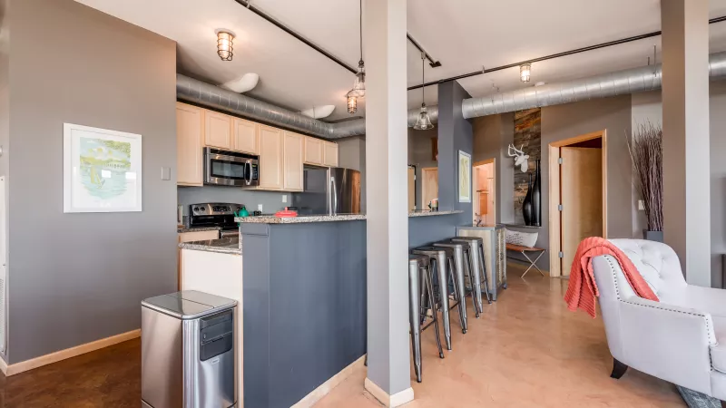 Open-plan industrial loft kitchen with light wood cabinets, dark gray island, and bar stools next to a living area.