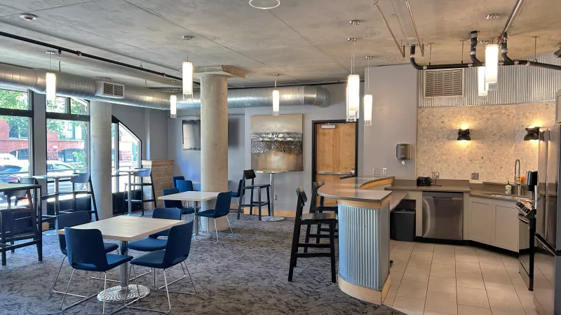 Modern common room with blue chairs, tables, and a kitchen area with stainless steel appliances under a concrete ceiling.