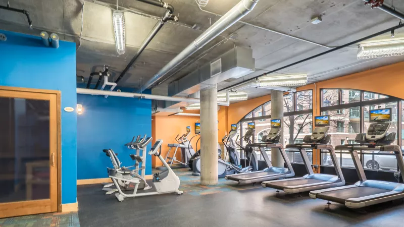 Modern gym with treadmills, ellipticals, blue and orange walls, exposed ducts, and large windows.