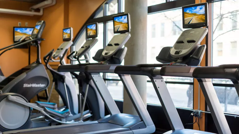 Modern gym with a row of treadmills, each with a screen, and an elliptical machine. Large windows, an orange wall.