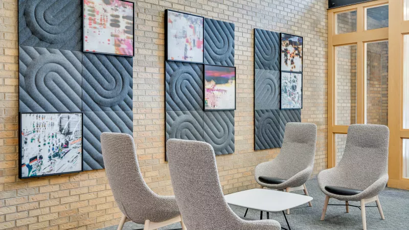 Modern lounge with three grey armchairs, a white coffee table, and framed abstract art on textured acoustic panels and brick.