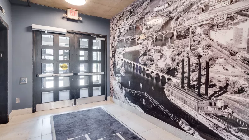 Modern hallway with a large black and white city mural, double glass doors, an elevator, and a dark rug with an "H" logo.