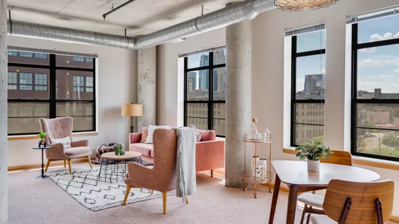 Modern loft living and dining room with pink furniture, concrete pillars, exposed ducts, and large windows showing city views.