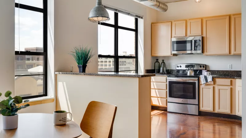 Modern apartment kitchen with light wood cabinets, stainless appliances, and a dining area by large windows.