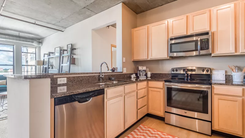 Modern kitchen with light wood cabinets, dark granite countertops, stainless steel appliances, concrete ceiling, and open layout.