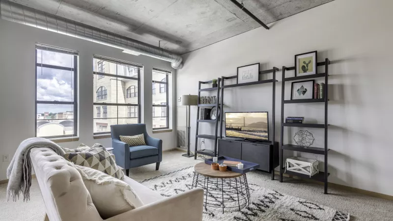 Modern living room with industrial ceiling, large windows, light couch, blue armchair, coffee table, and black shelves with TV.