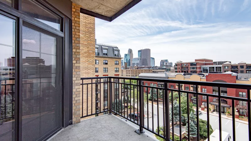 Apartment balcony with black railing, glass door, and a city skyline view under a bright blue sky with clouds.