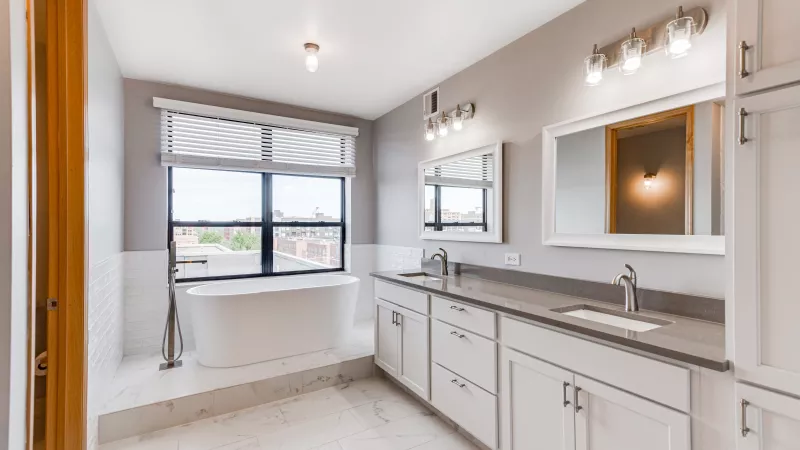 Modern bathroom with a white freestanding tub by a window, double vanity with gray counters and white cabinets. Photograph.