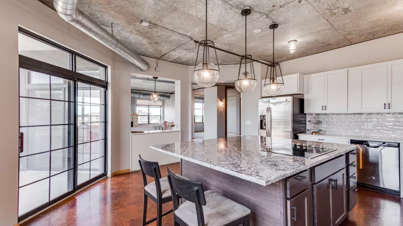 Modern kitchen with granite island, pendant lights, exposed concrete ceiling, dark wood floor, white cabinets, and glass sliding doors.