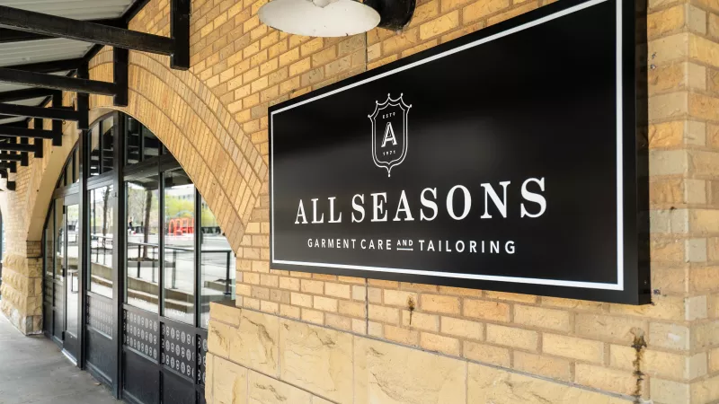 Black business sign for "All Seasons Garment Care and Tailoring" with a shield logo on a light brown brick wall.
