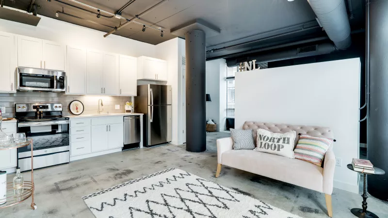 Modern open-plan studio with white kitchen, concrete floor, and light pink sofa on a geometric rug.