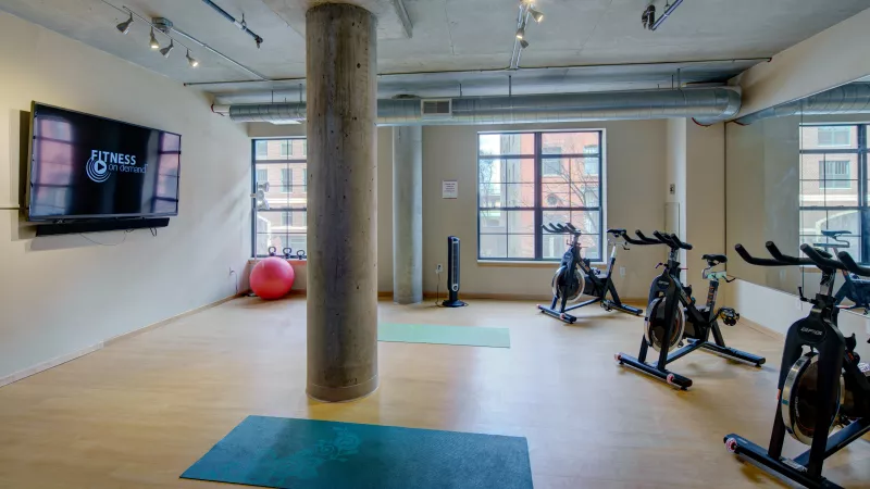 Modern gym with exercise bikes, yoga mats, a TV, and a red exercise ball. Concrete pillar in the center.