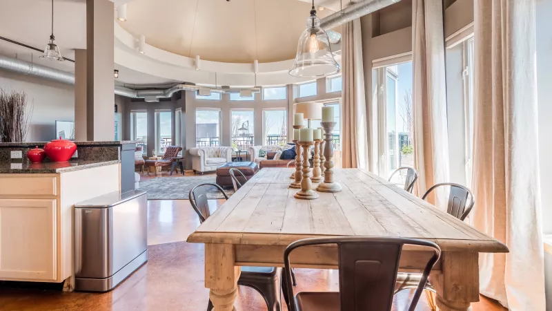 Bright, open-concept loft with a rustic dining table in the foreground, leading to a living area with large windows.
