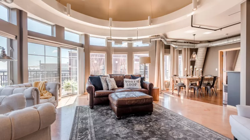 Lofty apartment living room with panoramic windows, brown leather couch, area rug, and dining space.