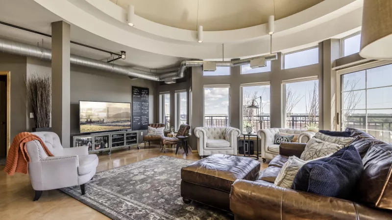 Modern loft living room with a curved window wall, leather sofa, multiple armchairs, large rug, and a TV.