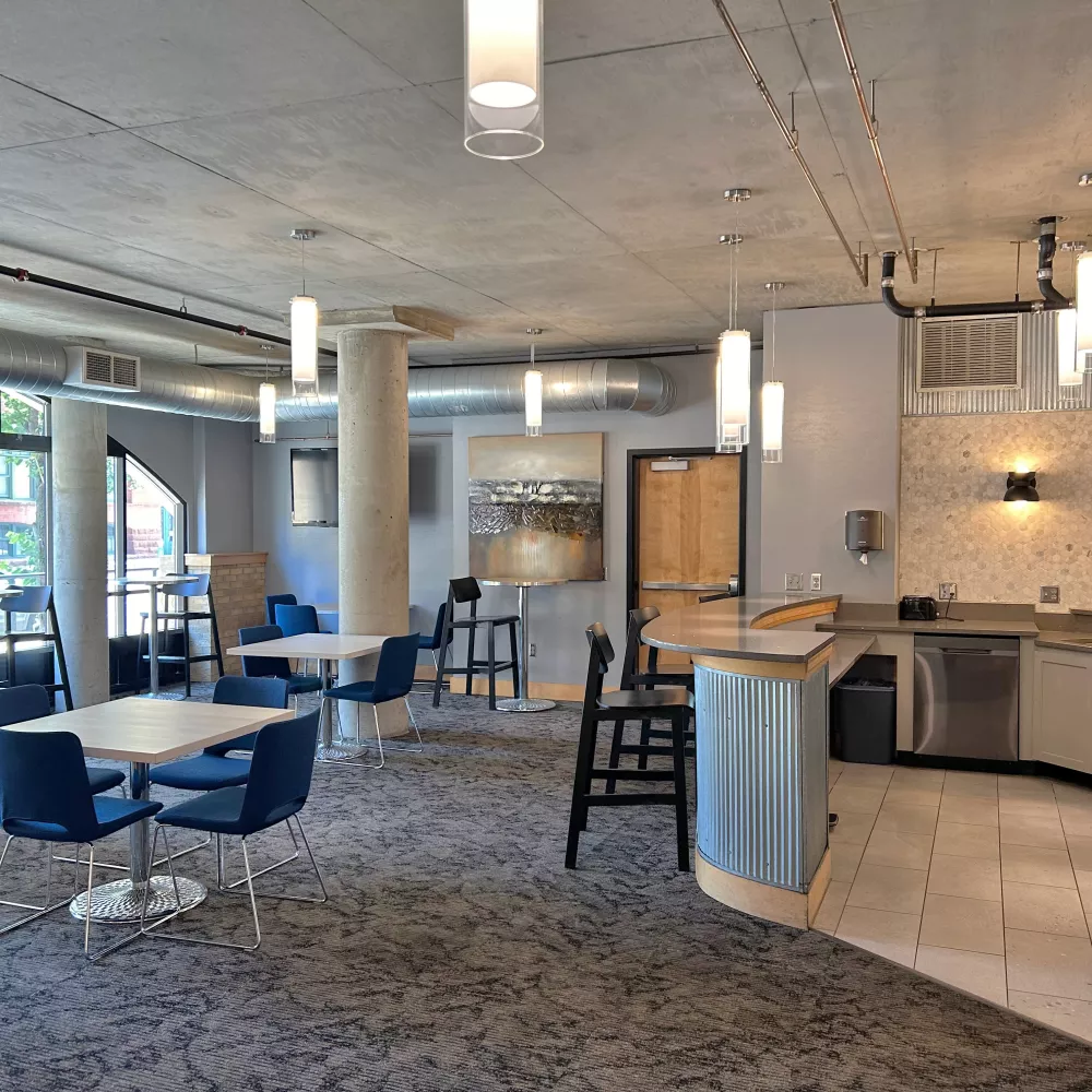 Modern common room with blue chairs, tables, and a kitchen area with stainless steel appliances under a concrete ceiling.