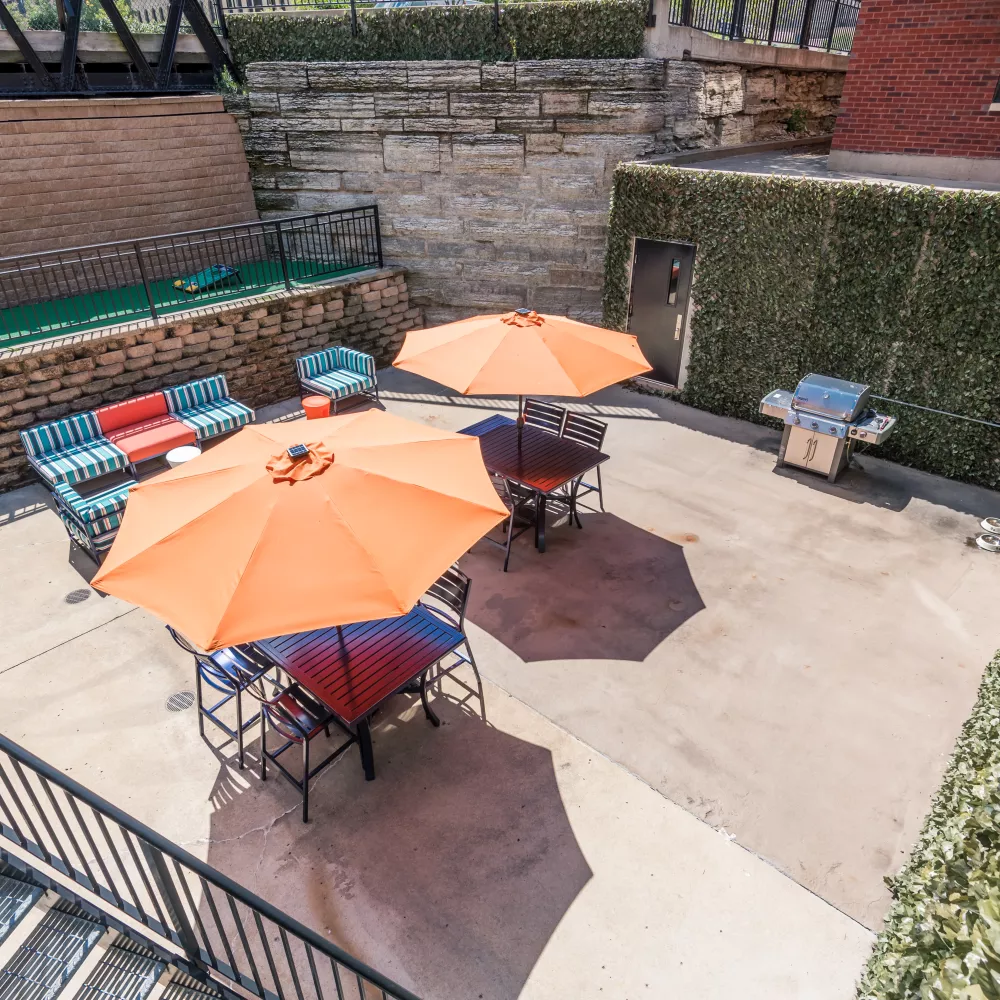 Outdoor patio with orange umbrellas, seating, and grills. Surrounded by brick, stone, and ivy walls.