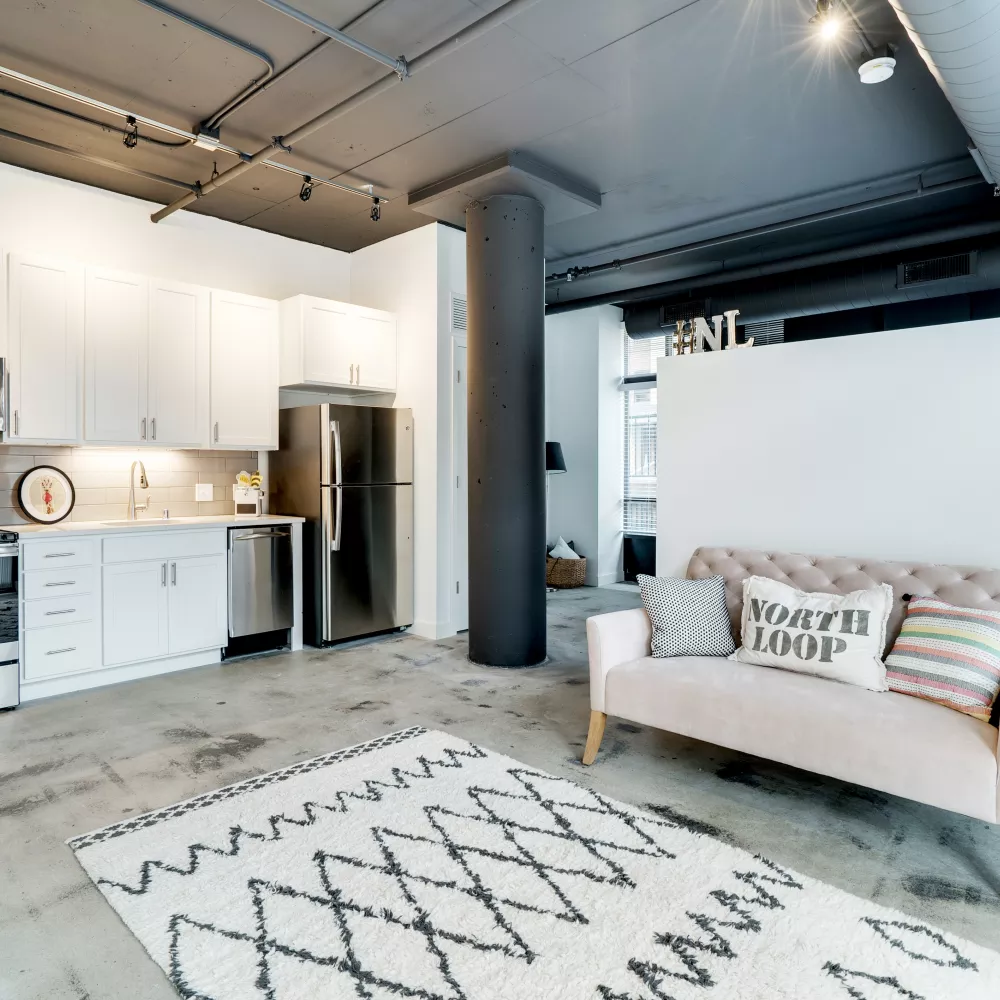 Modern open-plan studio with white kitchen, concrete floor, and light pink sofa on a geometric rug.
