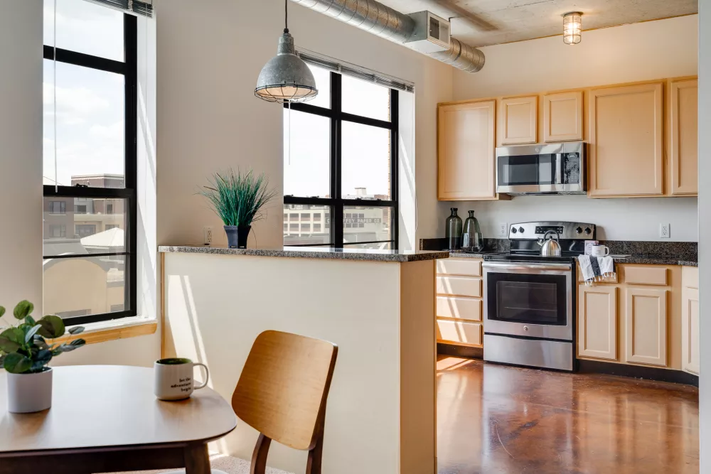 Modern apartment kitchen with light wood cabinets, stainless appliances, and a dining area by large windows.